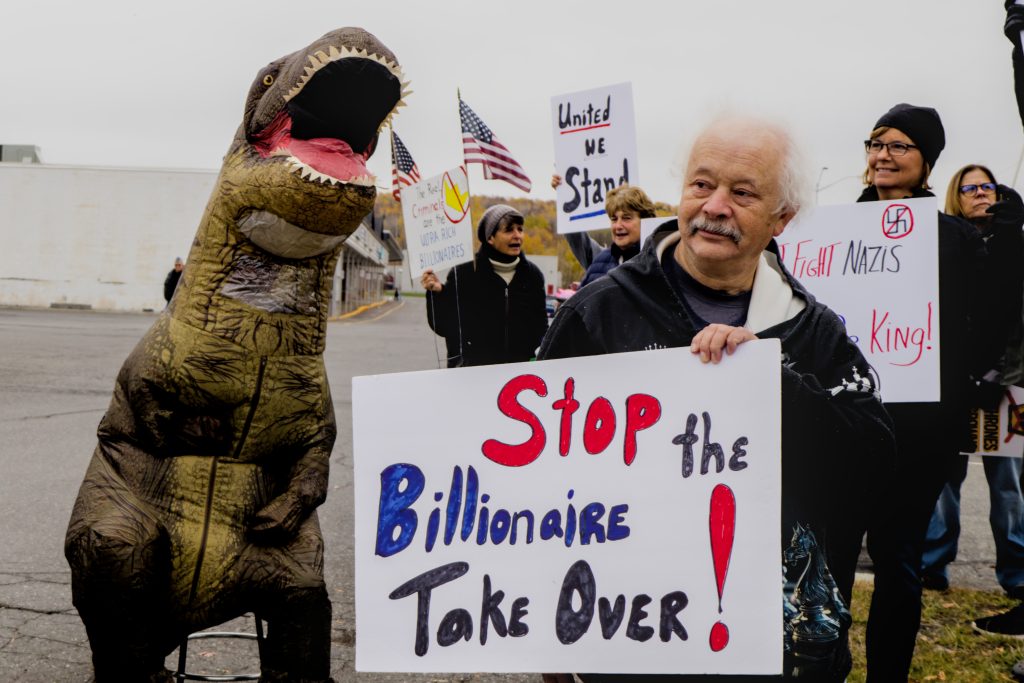 A person wearing an inflatable t-rex costume accompanies a man holding a sign that reads "Stop the Billionaire Take Over!" during the No Kings protest in Presque Isle, Maine, on October 18th, 2025.