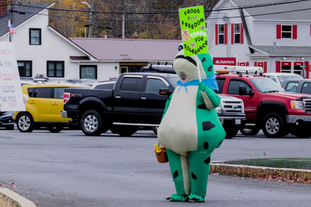 A person in an inflatable frog costume stands in the street holding a sign that reads "FROGS FOR FREEDON, TOADS FOR TOLERANCE" during the No Kings protest in Presque Isle, Maine, on October 18th, 2025.